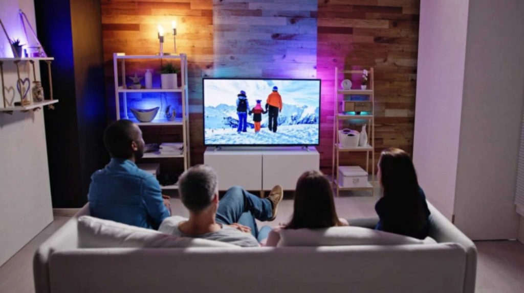 A family of four sitting on a white couch watching a winter scene on a large flat-screen TV in a cozy living room, with warm wood-paneled walls, modern shelving, and soft ambient lighting in blue and purple tones.